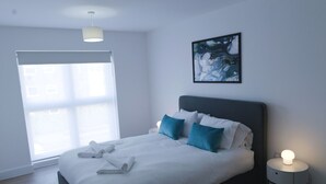 Ein modernes Schlafzimmer mit einem großen Bett, einem Nachttisch mit Lampe, einem Fenster mit Jalousien und einem Bild an der Wand.