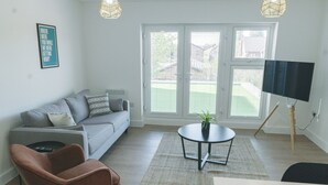 A modern living room with a grey sofa, a brown armchair, a small round table with a plant, and a TV on a stand. - Near Manchester Airport, Parking & EV Charger (Cheshire)