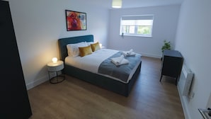 A bedroom with a bed, bedside table, lamp, artwork, window, and a small cabinet. - Near Manchester Airport, Parking & EV Charger (Cheshire)