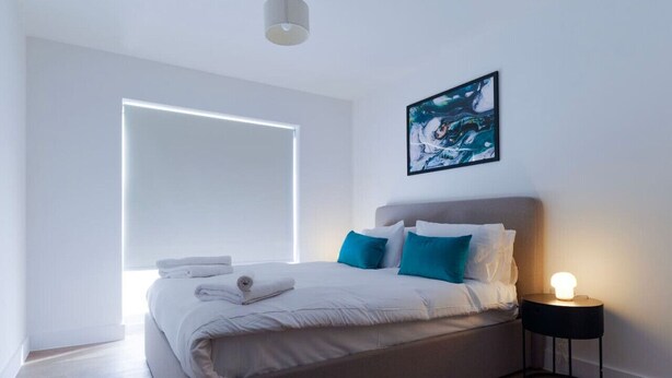A modern bedroom with a large bed, a side table with a lamp, a painting on the wall, and a window with blinds.