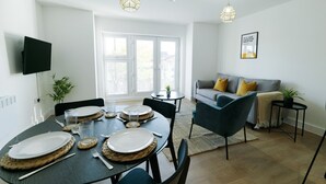 A modern living room with a dining area, a grey sofa with yellow cushions, and a flat-screen TV mounted on the wall.