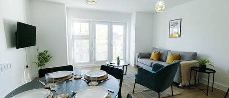 A modern living room with a dining area, a grey sofa with yellow cushions, and a flat-screen TV mounted on the wall.