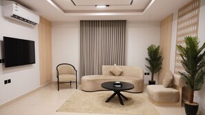 Een moderne woonkamer met een beige bank, een zwarte salontafel, een aan de muur bevestigde televisie en twee potplanten.