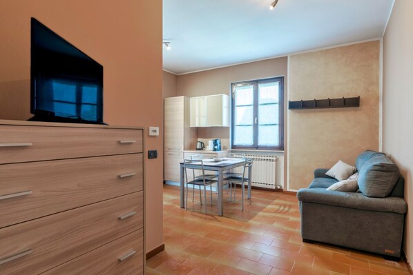 A modern living room with a sofa, a TV, a dining table with chairs, and a kitchen area with cabinets and a window.