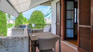 Un balcon avec une table et des chaises, une lampe, et une vue sur un arbre verdoyant et des bâtiments en arrière-plan.