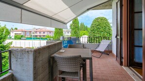 Un balcon avec une table et des chaises, une chaise avec un parasol, et une vue sur des bâtiments et de la verdure.