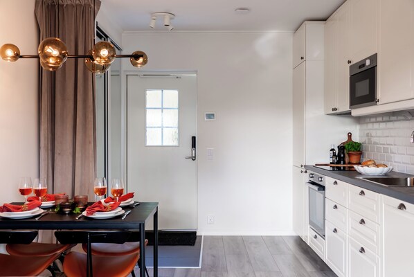 Une cuisine moderne avec des armoires blanches, un comptoir noir et un four encastré. On y trouve un coin repas avec une table dressée pour quatre personnes, un lustre et une fenêtre agrémentée de rideaux.