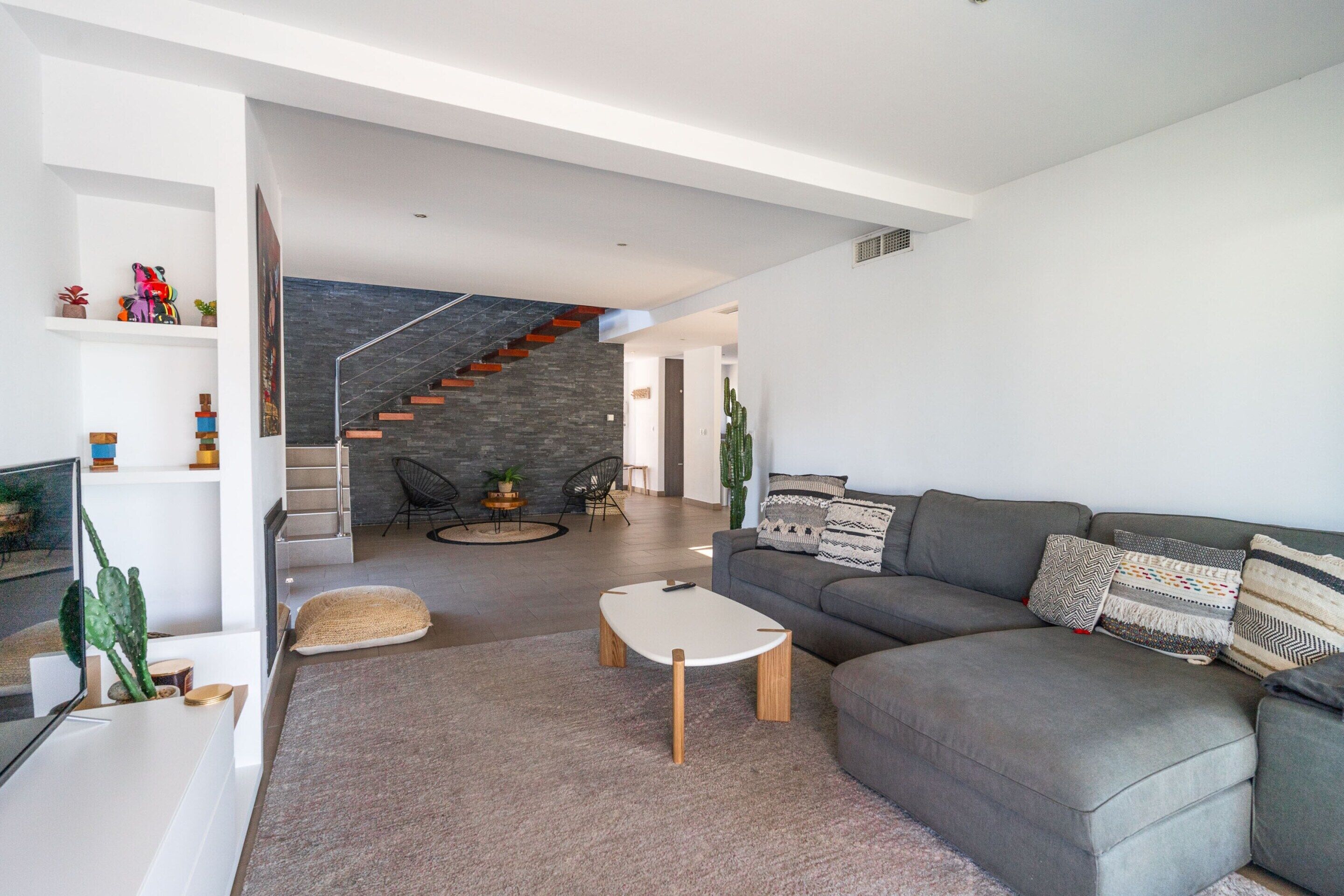 Una sala de estar moderna con un sofá gris, una mesa de centro blanca y una escalera con pared de piedra.