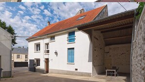 Un bâtiment blanc surmonté d’un toit de tuiles rouges, un balcon avec des volets bleus et un petit espace extérieur aménagé avec des sièges.