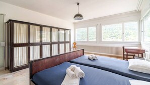A hotel room with two beds, a large wardrobe, a radiator, and a window with a view of greenery.