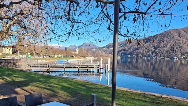 A lakeside dining area with a table and chairs, a dock, and a view of a mountain and buildings in the background.