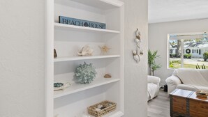 Una sala de estar con pared blanca, una estantería con objetos decorativos, un sofá blanco y una ventana que ofrece vistas a una casa y vegetación.