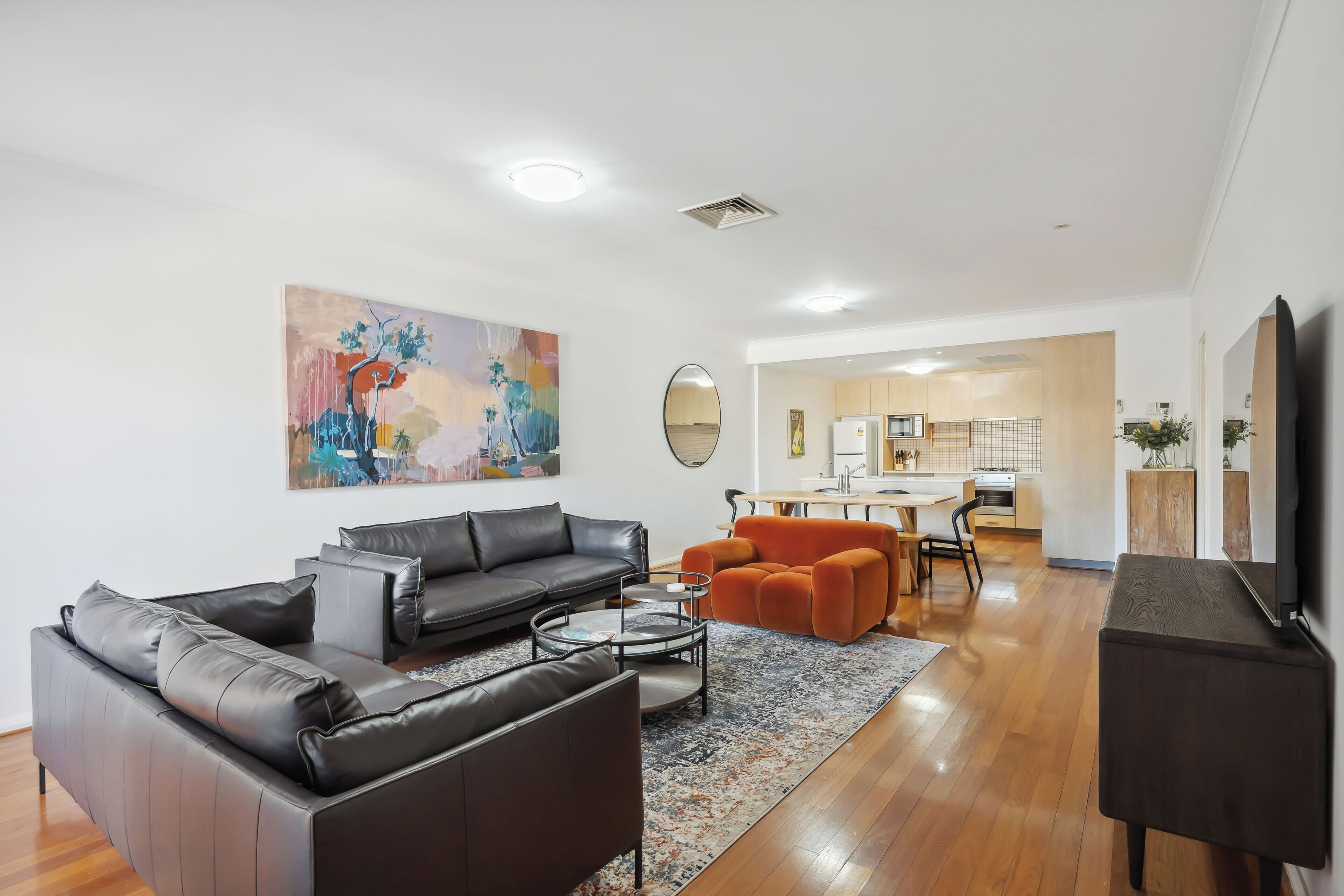 A modern living room with a black leather sofa, an orange armchair, and a coffee table. A large abstract painting adorns the wall.