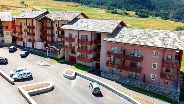 A multi-story hotel with balconies, a parking area, and a mountainous backdrop.