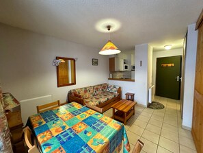 A small, cozy room with a wooden table, a colorful quilt-covered sofa, a small wooden bench, and a kitchen area with cabinets and a microwave. - Charming Termignon apartment for 4, 27m² with Wifi (Termignon)