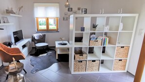 A modern living room with a leather chair, a coffee table, a white shelving unit with baskets, and a view through a window. - Modern Strata Montana vacation apartment with garden, underfloor heating and air conditioning (Bensheim)