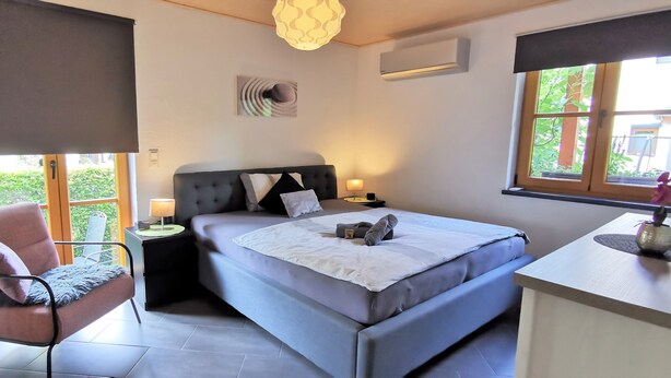 A bedroom with a bed, a chair, a nightstand, a lamp, and a window with a view of greenery. - Modern Strata Montana vacation apartment with garden, underfloor heating and air conditioning (Bensheim)
