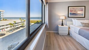 A hotel room with a large window offering a view of the beach and ocean. There is a bed with white bedding and a nightstand with a lamp. - Sandcastles 2BR/2BA Cocoa Beach Ocean Views! Heated Pool! (Cocoa Beach)