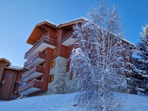 Un edificio de madera de varios pisos con detalles de piedra, un árbol cubierto de nieve al frente y un cielo azul despejado.