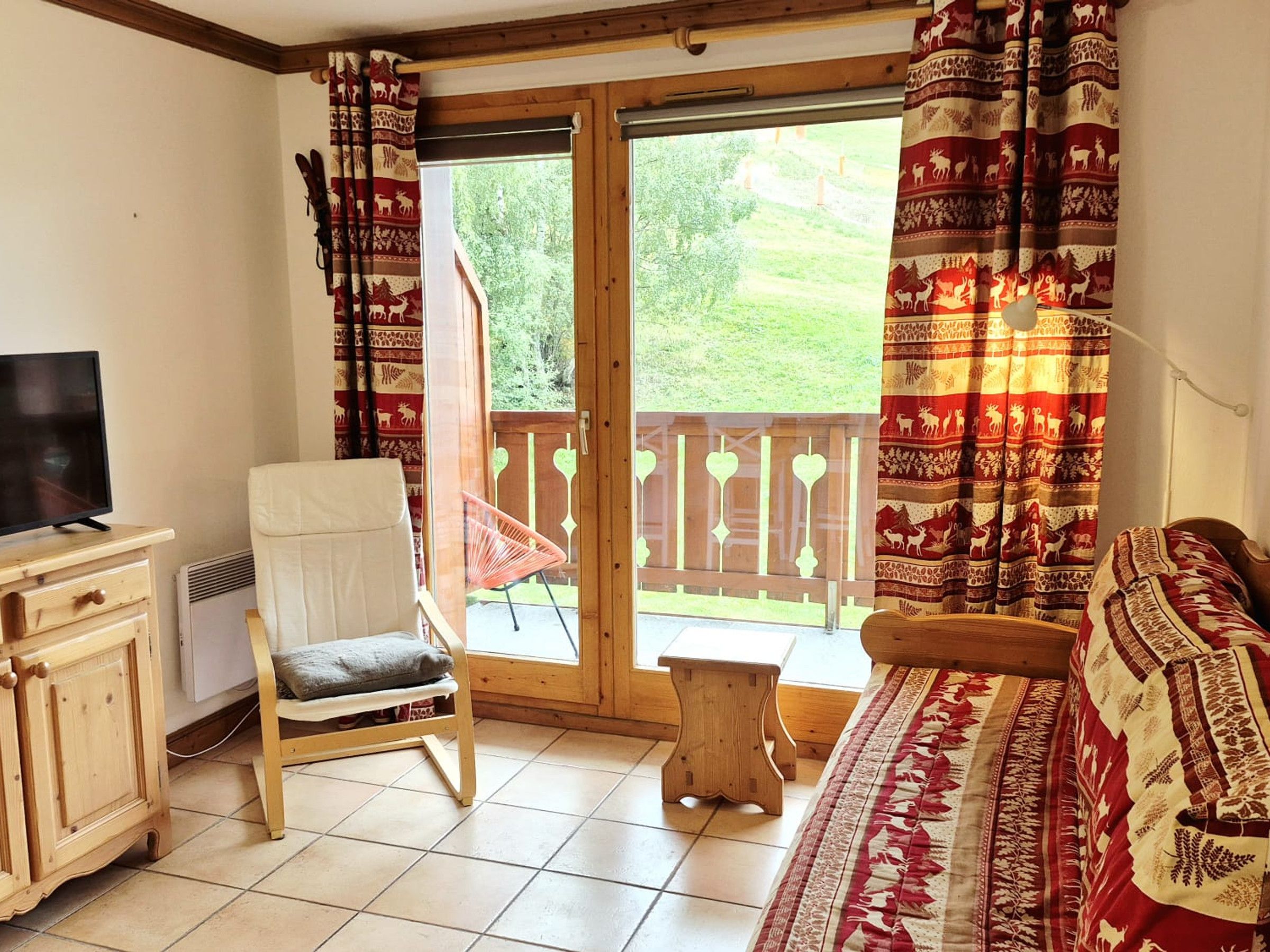 Habitación con televisión, una silla, un sofá y un balcón con cortinas estampadas.