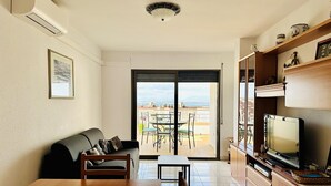 Een woonkamer met een bank, een eettafel en een televisie. Er is een balkon met uitzicht op de stad en de zee.