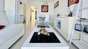 A modern living room with a white sofa, a dining table with chairs, a kitchen area with a white cabinet, and a black coffee table.