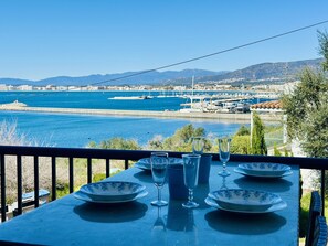 Uma mesa com pratos e copos, com vista para uma marina com barcos e um horizonte costeiro.