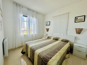 A bedroom with a bed, a window with curtains, a bedside table, a lamp, and two wall-mounted baskets.
