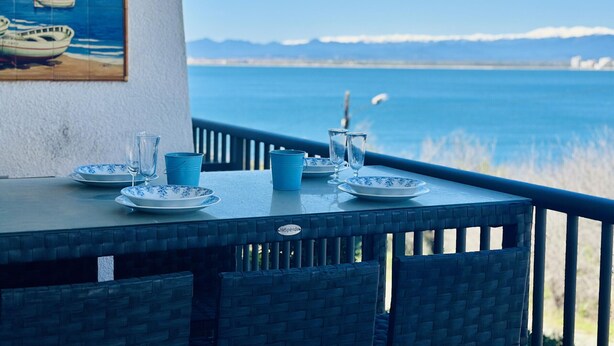 A table with plates, glasses, and a pitcher, overlooking a body of water with boats and mountains in the distance.