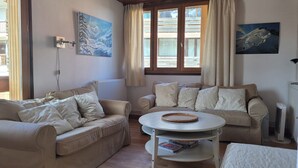 A cozy living room with two beige sofas, a round coffee table, and a radiator. There are two windows with curtains, a painting on the wall, and a lamp on a side table.