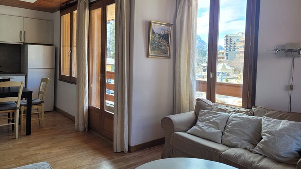 A cozy living room with a wooden ceiling, a beige sofa, a dining table with chairs, and large windows with curtains.