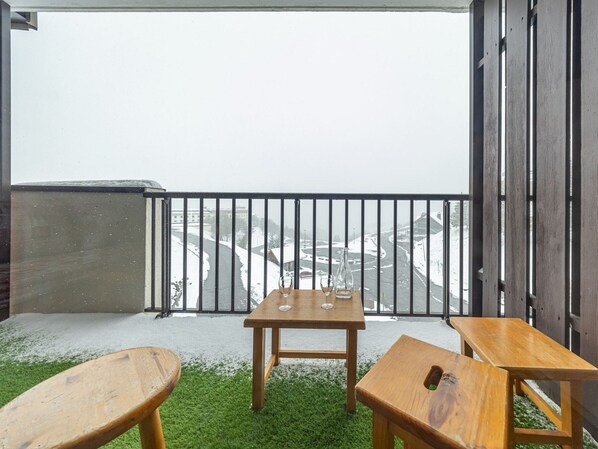 A balcony with a wooden table and two chairs, a metal railing, and a view of a snowy landscape.