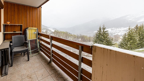 A balcony with a wooden railing, a chair, a small table, and a view of snow-covered mountains.