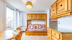 A compact room with a wooden bunk bed, a dining table with chairs, a kitchen area with wooden cabinets, and a hanging light fixture.