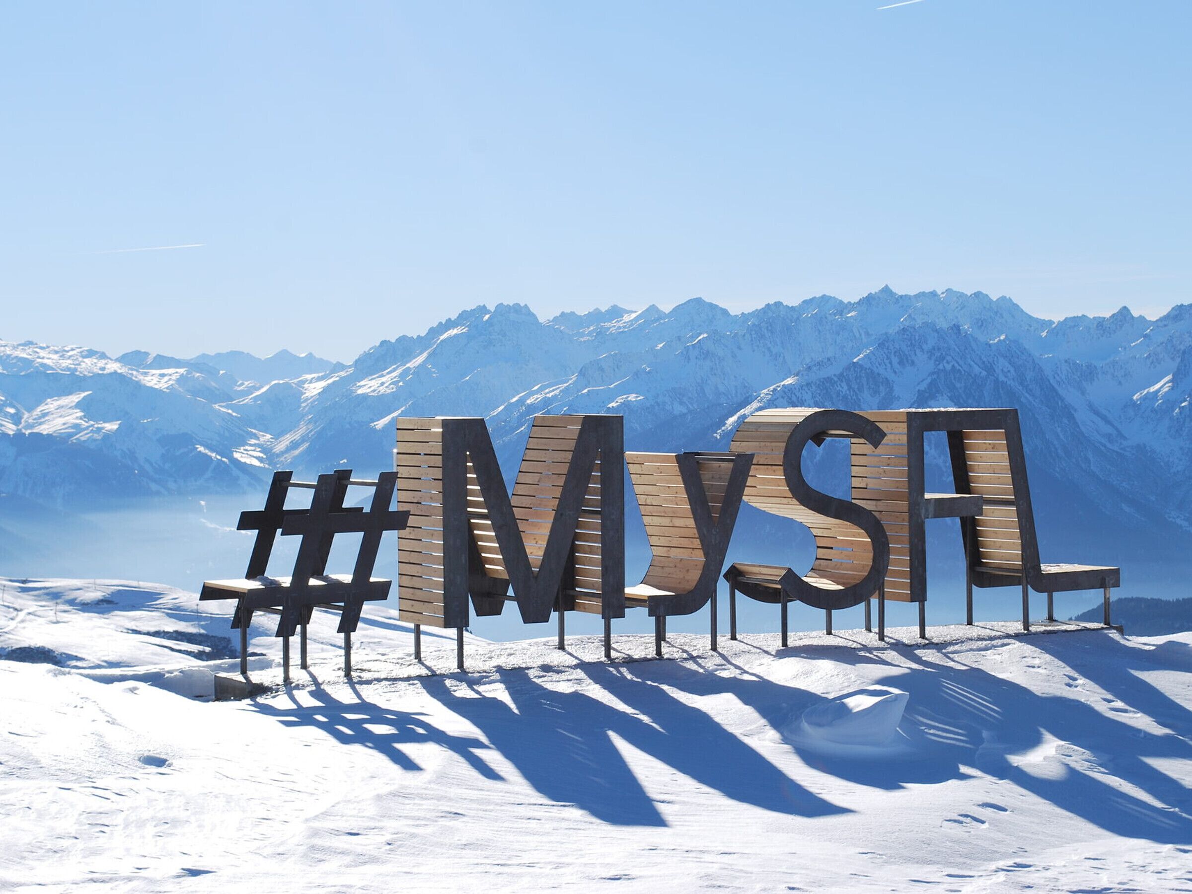 A wooden bench with the letters "MYS" and hashtags "#MYS" in the foreground, set against a backdrop of snow-covered mountains and clear blue sky.