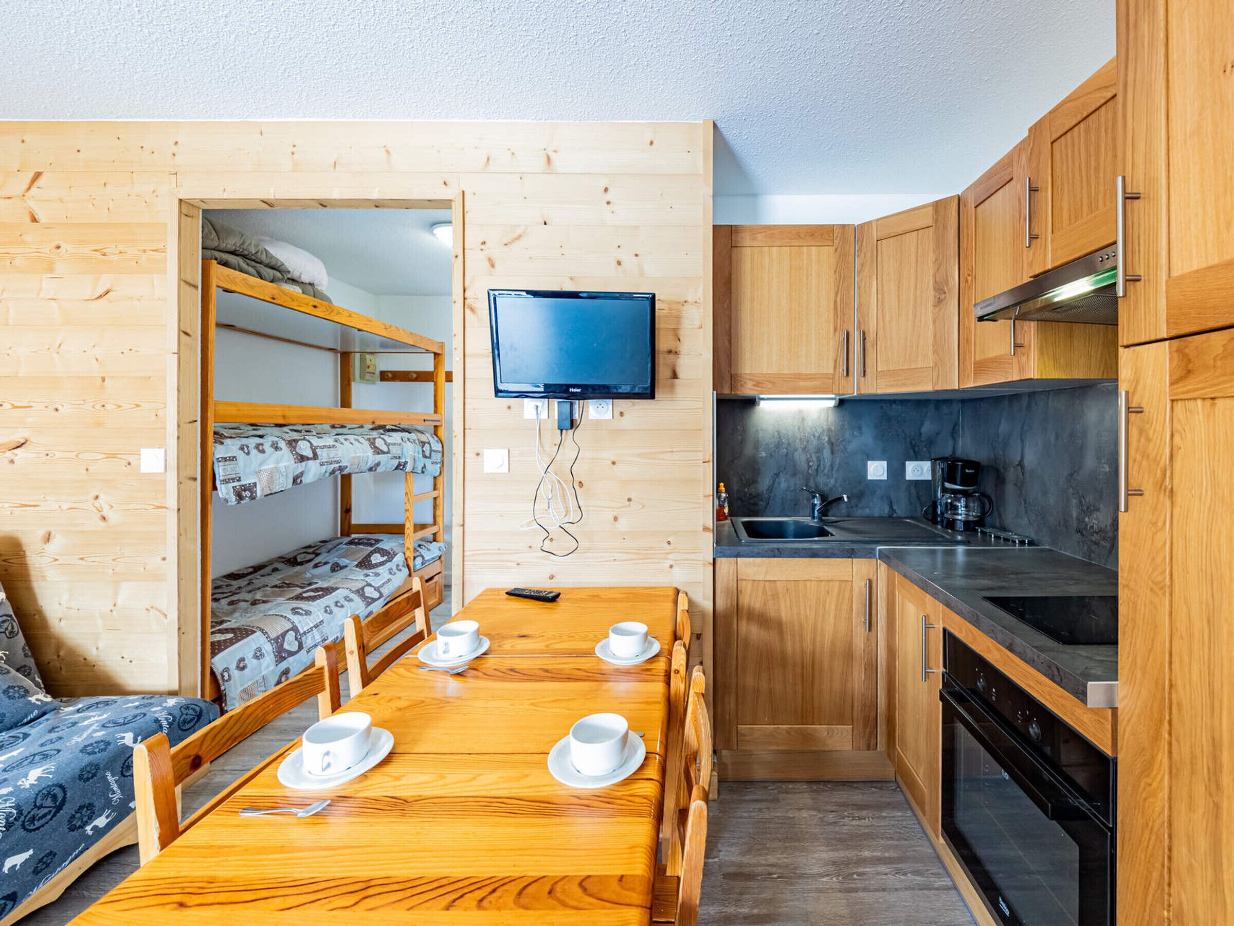 Una habitación compacta con literas, una mesa de comedor con tazas, un televisor y una kitchenette con estufa y pileta.