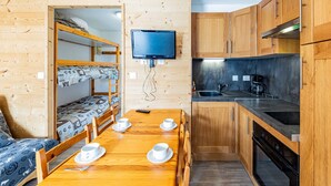Een kleine kamer met een stapelbed, een eettafel met kopjes, een televisie en een kitchenette met een kookplaat en wastafel.