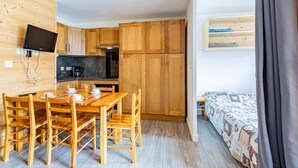 Een knus ingerichte kamer met een houten eettafel en stoelen, een kitchenette, een bed met een bedovertrek met dessin en een wandgemonteerde televisie.