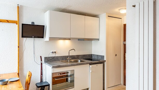 Una cocina compacta con armarios blancos, horno integrado, lavamanos y televisor montado en la pared.