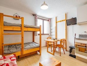 A dormitory room with bunk beds, a dining table with chairs, a TV mounted on the wall, and a kitchenette area. - Charming 19m² studio for 4 in Saint-François-Longchamp (Saint-François-Longchamp)