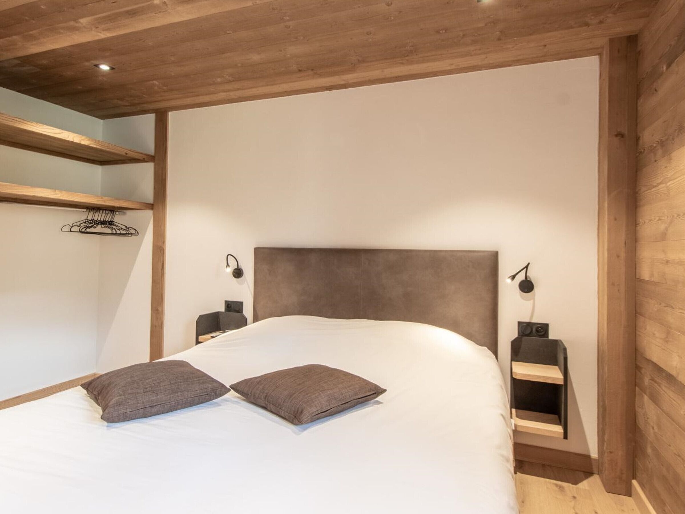 A bedroom with a wooden headboard, two bedside tables with lamps, a bed with white bedding and two pillows, and a wooden shelf unit.