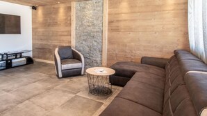 Ein modernes Wohnzimmer mit Holzbalkendecke, einer Wand aus Stein, einem braunen Ledersofa, einem gepolsterten Sessel und einem kleinen runden Couchtisch.