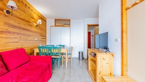 Un espacio habitable pequeño con un sofá rojo, sillas color turquesa, una mesa de comedor de madera y un televisor sobre un mueble de madera.