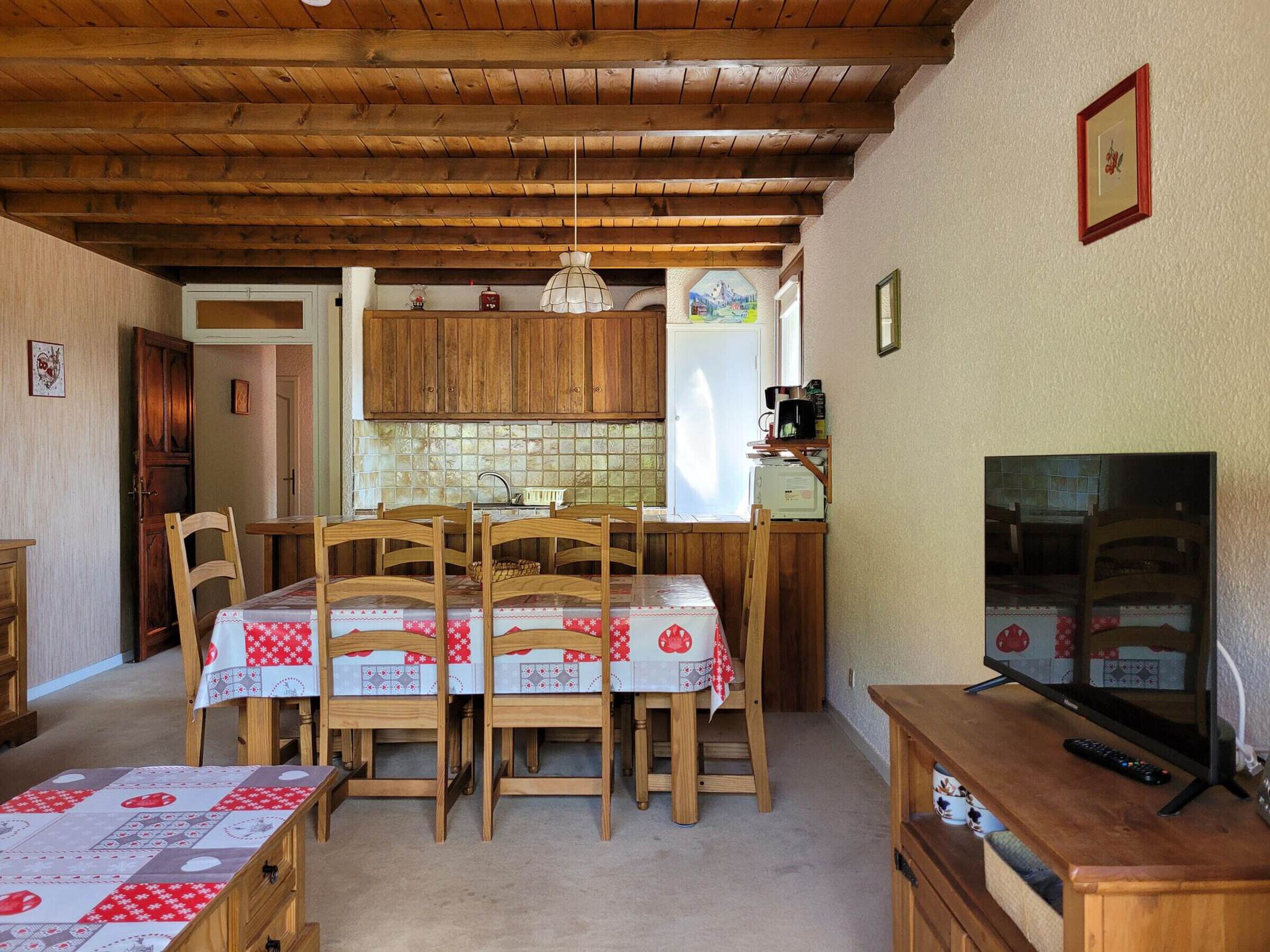 A cozy kitchen with wooden cabinets, a dining table with chairs, a television, and a refrigerator.