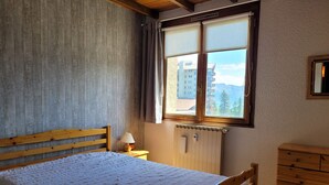 A bedroom with a wooden bed, a nightstand with a lamp, a dresser, and a window with a view of buildings and mountains. - 3 rooms for 6, south/east balcony near slopes (Pra-Loup)