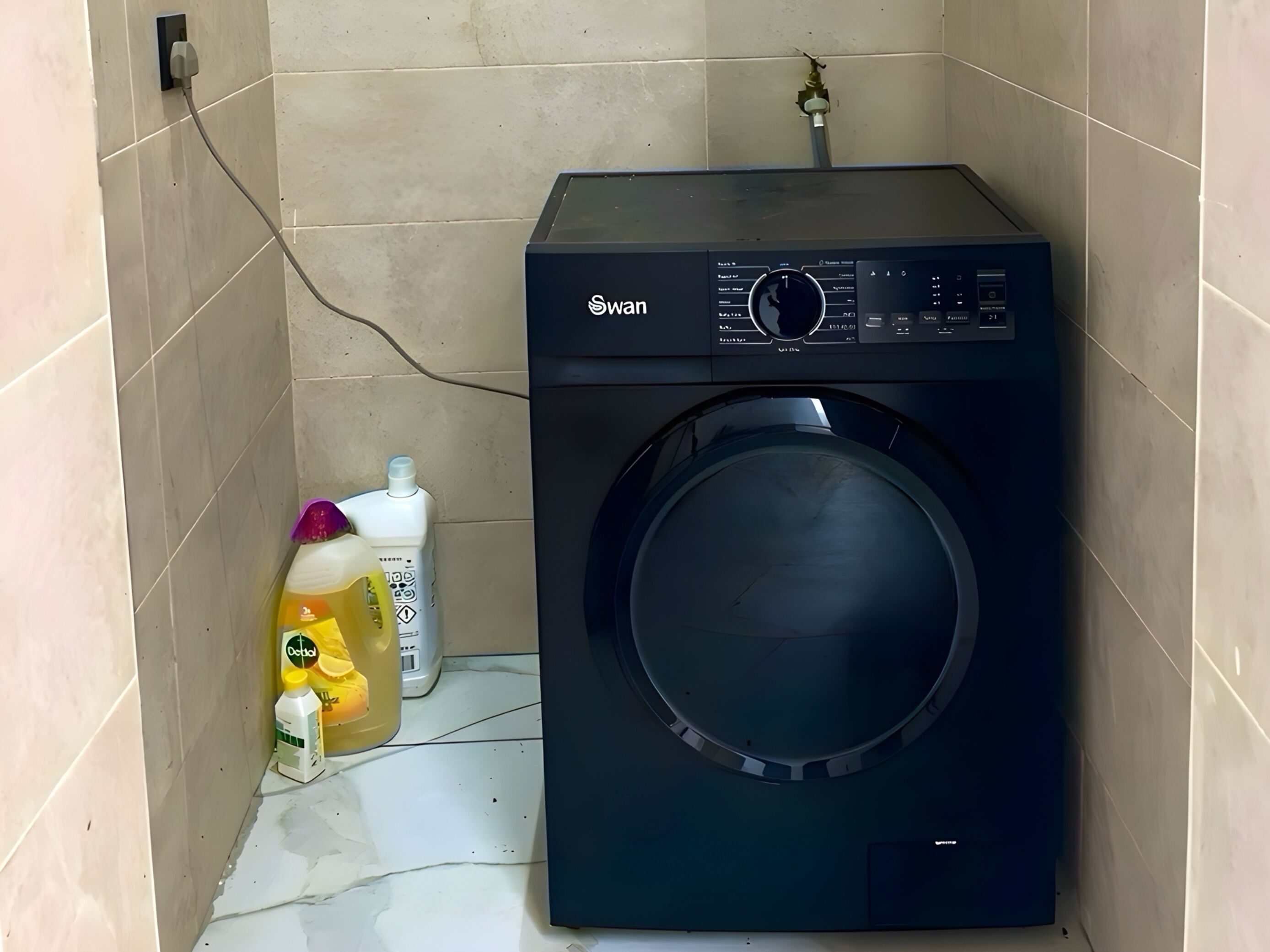 A compact, black front-loading washing machine with a control panel, situated in a small, tiled laundry room.