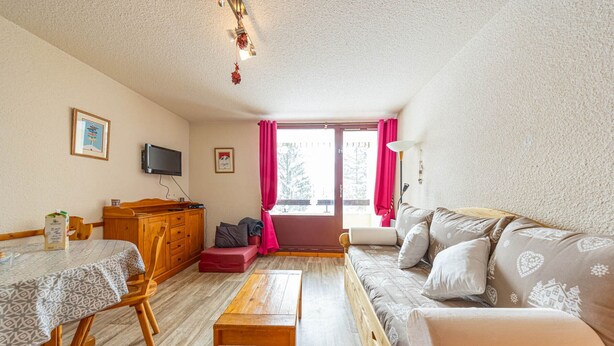 Una habitación acogedora con un sofá de madera, una mesa de comedor con sillas, una mesita de madera, un televisor en la pared y una ventana con cortinas.
