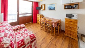 A cozy room with a wooden floor, a red and white quilted bed, a dining table with chairs, a microwave, a small cabinet, and a window with red curtains.