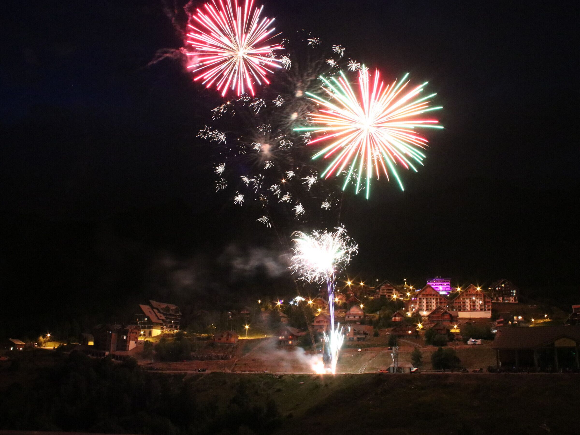 Vista nocturna de un pueblo con edificios y un espectáculo de fuegos artificiales.