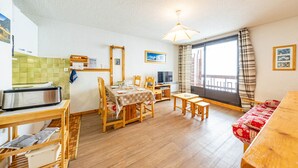 A cozy living room with a dining area, wooden furniture, a kitchen with a toaster, and a large window with curtains.